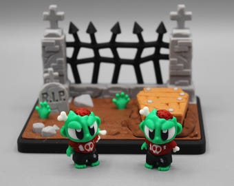 May include: A miniature Halloween-themed diorama featuring two green zombie figures with exposed brains and bones. The scene includes a graveyard with a tombstone, a coffin, and a black fence. The figures are wearing red shirts and black pants.