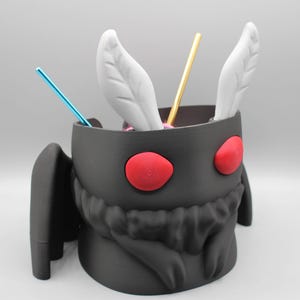 May include: A black, stylized container shaped like a moth with red eyes and white feather-like accents. The container holds blue and gold straws. The container has two black wings.