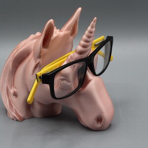May include: A pink unicorn head-shaped glasses holder with a horn and ears. The holder has black-framed glasses with yellow arms resting on the unicorn's horn. The glasses holder is a decorative item.