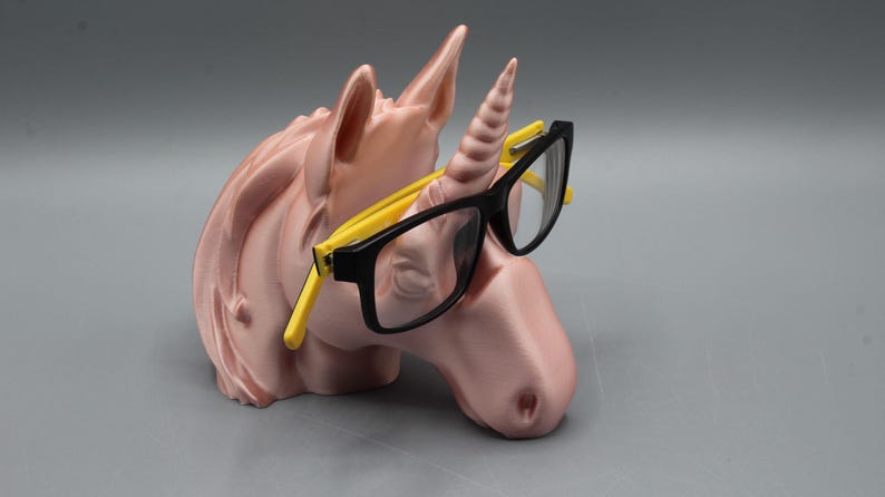 May include: A pink unicorn head-shaped glasses holder with a horn and ears. The holder has black-framed glasses with yellow arms resting on the unicorn's horn. The glasses holder is a decorative item.