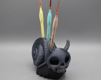 May include: A dark gray, 3D-printed pen holder shaped like a snail with a skull head and horns. Three paint brushes with yellow, teal, and beige handles are inserted into the holder. The holder sits on a black base.