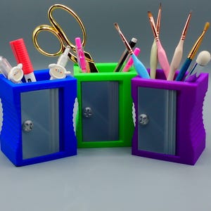 May include: Three colorful pencil sharpener-shaped containers, blue, green, and purple, each holding various art supplies. The blue container holds a red marker and white objects. The green container has gold scissors. The purple container has paint brushes.