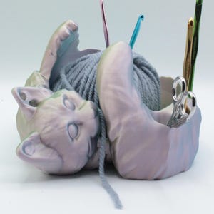 Puede incluir: Un bol de lana de cerámica con forma de gato, con una bola de lana gris en su interior. El gato está tumbado de lado con la cabeza apoyada en la lana. Hay tres agujas de crochet y un par de tijeras en el bol.