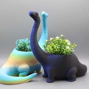 Puede incluir: Tres macetas con forma de dinosaurio, una azul, una verde y una negra, con plantas que crecen en ellas.
