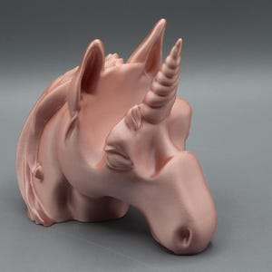 May include: A rose gold unicorn head sculpture with a detailed horn, ears, and mane. The sculpture is a decorative item, possibly a figurine or a holder, and is set against a gray background.