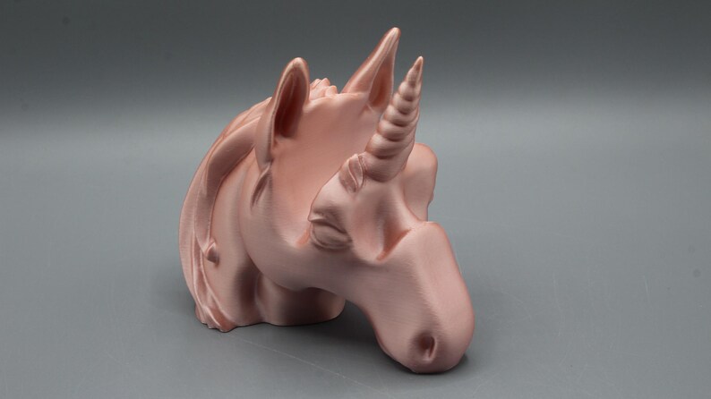 May include: A rose gold unicorn head sculpture with a detailed horn, ears, and mane. The sculpture is a decorative item, possibly a figurine or a holder, and is set against a gray background.