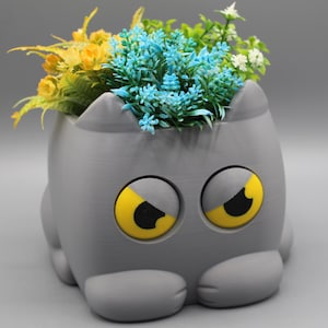 May include: A gray cat-shaped planter with large, yellow, downward-sloping eyes and small ears. The planter is filled with a variety of artificial flowers in shades of yellow, light blue, and white, which sprout from the top of the cat's head.