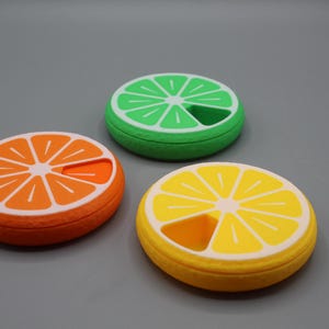 Puede incluir: Tres pastilleros circulares de colores, diseñados para parecerse a rodajas de cítricos: naranja, lima y limón. Cada estuche tiene un borde blanco y una sección recortada. Los estuches están sobre una superficie gris. Medidas en cm.