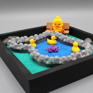 May include: A black framed miniature zen garden with a blue pond, gray rocks, a purple flower, and four yellow rubber duckies. A yellow chick figurine sits on a wooden bench.