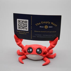 May include: A red crab-shaped card holder with large, cartoonish eyes and a white belly. The crab holds a navy blue card with a QR code and the text "The Empty Nest Since 2021". The card also states "Veteran owned and operated".