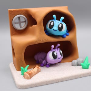 May include: A brown, two-story tree-shaped structure with a circular window, housing a blue insect on the top level and a purple insect on the bottom level. The base is light-colored with a small brown log, two green grass tufts, and two gray stones.