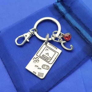May include: A silver keychain featuring a Game Boy-style charm with the word "Game Boy" printed on it. The keychain also includes a silver letter "C" charm and a red rhinestone charm.