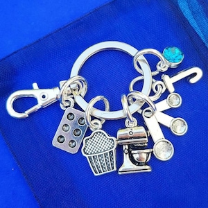 May include: Silver keyring with a variety of baking-themed charms, including a cupcake, a mixer, a measuring spoon, and a baking sheet. The keyring also features a blue gemstone and a letter "J".