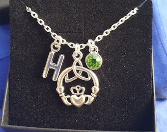 Pode incluir: Colar de prata com um pingente de cristal verde, um pingente de nó celta e um pingente com a letra H.