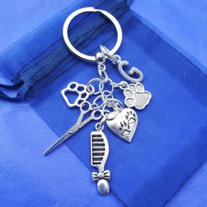 May include: Silver charm keyring with a paw print, scissors, comb, heart with paw prints, and the letter G. The charms are attached to a silver key ring.