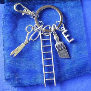 May include: A silver keychain with a ladder, paintbrush, scissors, and the letter "L" charm. The keychain is hanging on a blue mesh bag.