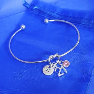 May include: Silver bangle bracelet with a knot charm, a charm with the letter "M" and a charm with the number "21". A pink rhinestone charm is also attached to the bracelet.