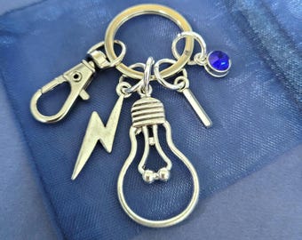 May include: A silver-toned metal keychain featuring a circular ring, a lobster clasp, a lightning bolt charm, a light bulb charm, a small rectangular bar charm, and a single round blue rhinestone charm attached to the main ring.