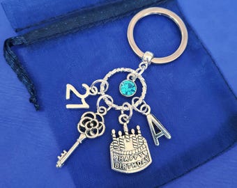 May include: Silver keychain with a teal blue crystal charm, a key charm, a cake charm with the text "Happy Birthday", and a letter "A" charm. The keychain is attached to a silver ring.