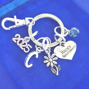 Puede incluir: Llavero de plata con dijes que incluyen una flor, un coraz&oacute;n con el texto "Happy Retirement", una letra "C", el a&ntilde;o "2023" y un cristal azul.