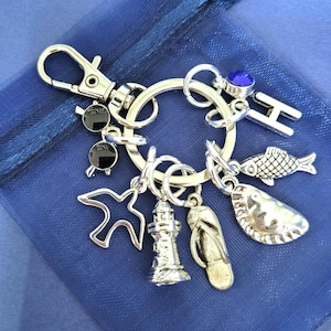 May include: A silver-toned metal keychain featuring a circular ring and a lobster clasp. Attached charms include a lighthouse, a flip-flop, a fish, a bird silhouette, a pair of black sunglasses, a letter H, and a small round blue crystal bead.