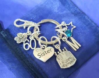 May include: Silver keychain with charms including a heart with the word "Sister", a cake with the words "Happy Birthday", a star, a flower, a number 60, and the letter "M".