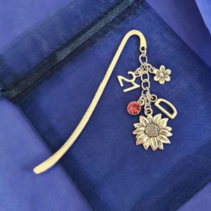 May include: A gold-colored metal bookmark with a curved top. It features a dangling charm cluster with a sunflower, a red gem, the number 21, a flower, and the letter D. The bookmark rests on a dark blue fabric.