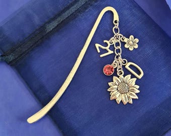 May include: A gold-colored metal bookmark with a curved top. It features a dangling charm cluster with a sunflower, a red gem, the number 21, a flower, and the letter D. The bookmark rests on a dark blue fabric.