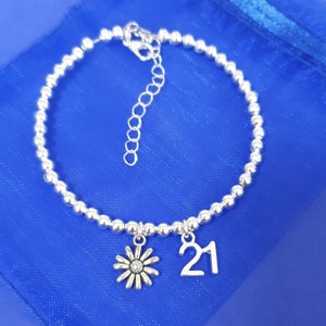 May include: A silver beaded bracelet with a silver daisy charm and a silver charm with the number 21 on it.