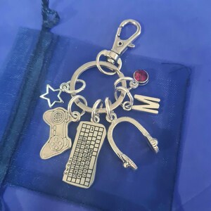 May include: Silver-toned keychain with multiple charms, including a star, a game controller, a keyboard with letters and numbers, a headset, and the letter "M". A red gem is also attached. The keychain is on a blue background.