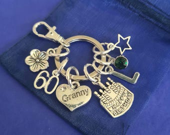 May include: Silver-toned keychain with multiple charms, including a flower, the number 60, a heart with "Granny" engraved, a star, a green gem, and a cake with "Happy Birthday" text. The keychain is on a blue pouch.