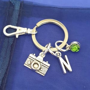 May include: Silver keyring with a camera charm, a letter N charm, and a green gemstone. The keyring has a silver split ring and a silver lobster clasp.