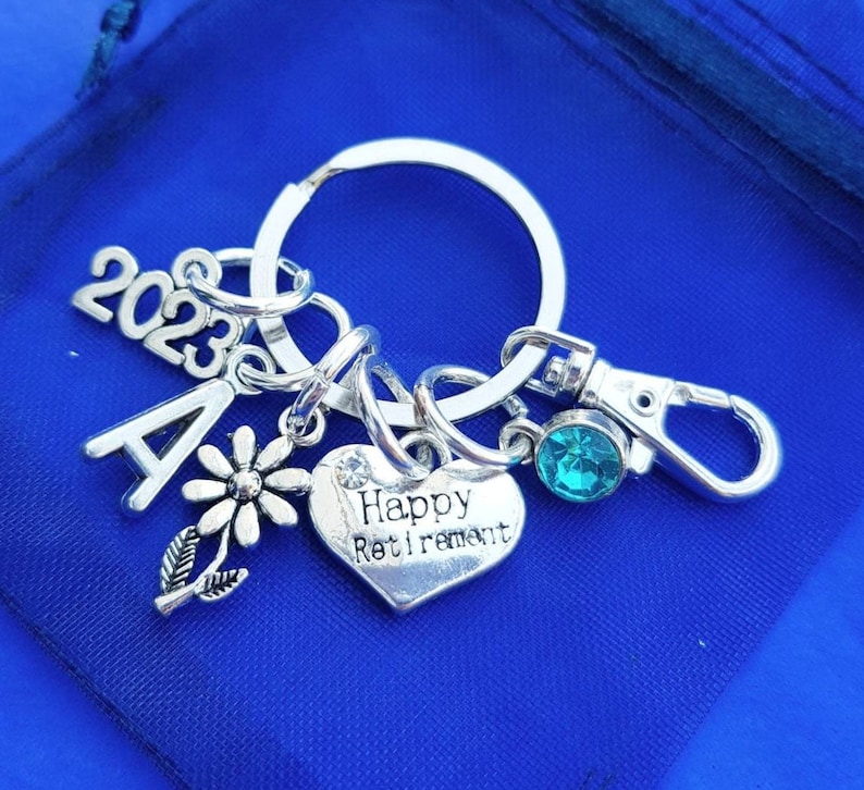 Puede incluir: Llavero de plata con abalorios, incluyendo un abalorio en forma de coraz&oacute;n con el texto "Happy Retirement", un abalorio de flor, un abalorio con la letra "A" y un abalorio 2023. El llavero tambi&eacute;n tiene una gema azul turquesa y un cierre de plata.