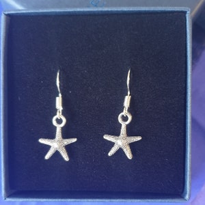 May include: A pair of silver starfish earrings presented in a blue box. The earrings feature a textured starfish design, suspended from silver hooks. The box has a dark blue interior and a blue exterior.