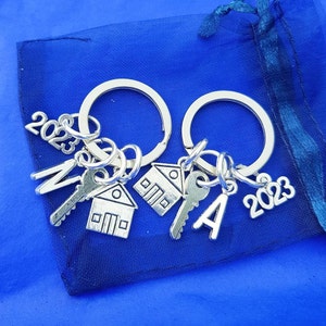 May include: Two silver keyrings with house, key, and letter charms. The charms are attached to the keyrings with silver rings. The keyrings are on a blue background. The charms have the numbers 2023 on them.