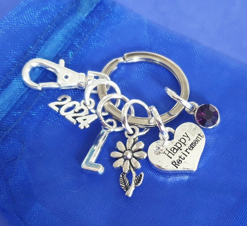Puede incluir: Llavero de plata con un dije en forma de coraz&oacute;n que dice "Happy Retirement" y un dije de cristal morado. El llavero tambi&eacute;n tiene un dije de plata con el n&uacute;mero 2024, un dije de plata con la letra L y un dije de plata con una flor.