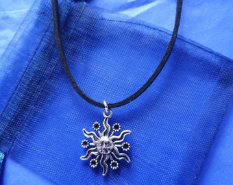 May include: A silver sun pendant necklace on a black suede cord. The pendant features a stylized sun with a face and rays. The necklace is displayed on a blue mesh bag.