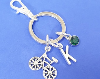May include: Silver keychain with a small bicycle charm, a letter K charm, and a green gemstone charm.