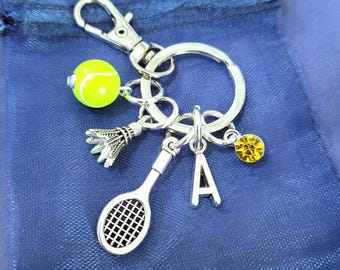 May include: A silver-toned metal keychain featuring a circular ring and a lobster clasp. Attached charms include a bright neon yellow tennis ball, a silver badminton shuttlecock, a silver tennis racket, a silver letter A, and a small yellow crystal.