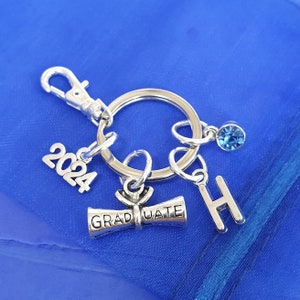 May include: Silver graduation keyring with a blue charm, a diploma charm with the word "GRADUATE" on it, a charm with the year "2024" on it, and a charm with the letter "H" on it.