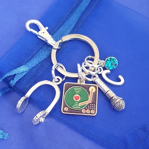 May include: Silver keychain with a record player charm, headphones charm, microphone charm, a letter C charm, and a green gem.