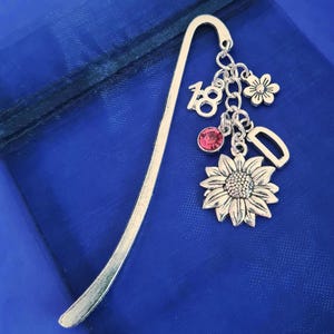May include: Silver bookmark with a curved top and dangling charms. Charms include a sunflower, a pink gem, the number 18, a small flower, and the letter D. The bookmark rests on a blue fabric surface.