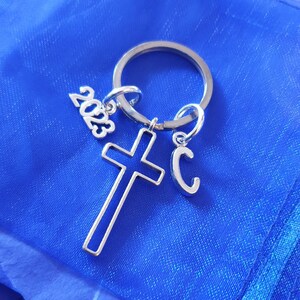 May include: Silver keychain with a cross charm, a letter 'C' charm, and a 2023 charm. The keychain is on a blue background.