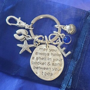 May include: Silver keyring with a charm that reads "may you always have a shell in your pocket & sand between your toes". The keyring also features a starfish, a seashell, a blue crystal, and a small charm of two footprints.