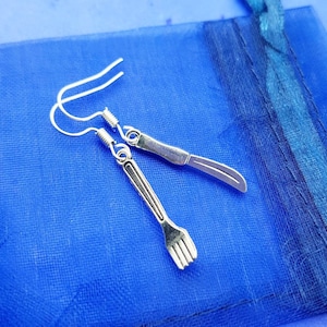 May include: A pair of silver earrings shaped like a knife and fork. The earrings are hanging from a blue background.
