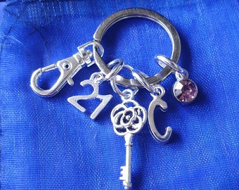 May include: Silver keychain with a key charm, a rose charm, a number 21 charm, a letter C charm, and a small purple rhinestone charm. The keychain is on a blue background.