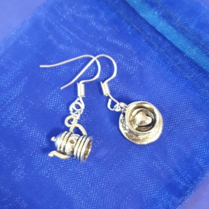 May include: A pair of silver earrings with a teapot and a teacup charm. The teapot charm has a spout and a handle. The teacup charm has a heart-shaped handle.
