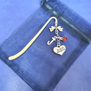 May include: Silver bookmark with a heart charm that says "Grand Daughter" and a red crystal charm. The bookmark is in a blue organza bag.