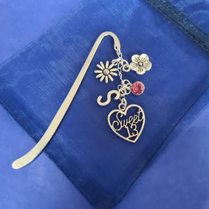 May include: Silver metal bookmark with a daisy and flower charm, a red gem, and a heart pendant that says "Sweet 13". The bookmark is on a blue fabric background.