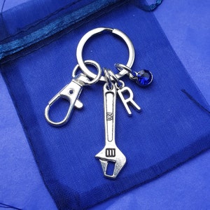 May include: Silver-toned keychain with a wrench charm, letter "R", and a blue crystal accent. The keychain includes a clasp and a key ring. The background is a blue fabric.